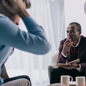 Frustrated young man sititng on sofa and african american psychiatrist in office