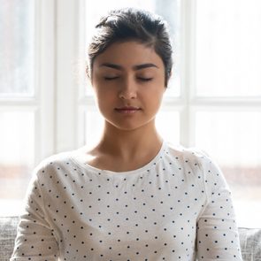 Calm Indian woman with closed eyes meditating at home alone close up, relaxed beautiful girl sitting in lotus pose on comfortable couch, dreaming, doing yoga exercise, stress relief concept