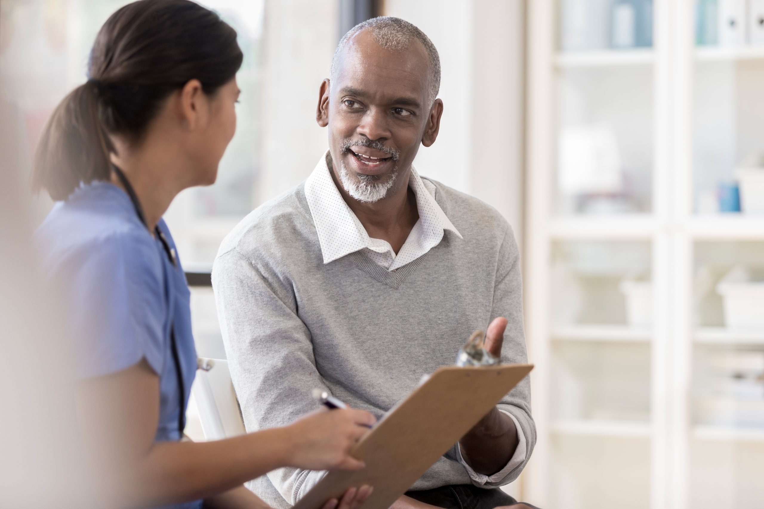 A cheerful senior man sits next to an unrecognizable female doctor in her office. She holds a clipboard as he asks questions regarding his diagnosis.