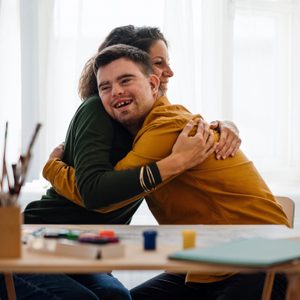 Young man with Down syndrome sitting and embracing with his tutor after painting indoors at home, homeschooling.